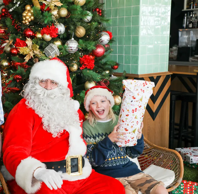 Santa with a smiling child holding a wrapped gift beside the Christmas tree